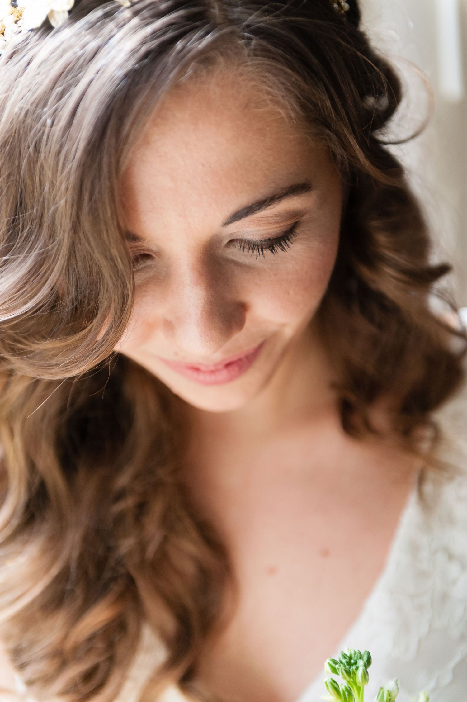 A bride with soft curled brown hair and a floral crown smiles gently downward, wearing a lace dress.