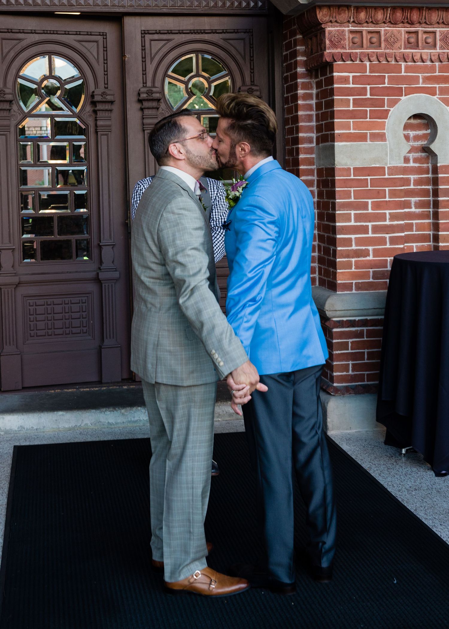 Two grooms in suits share a kiss outside a brick building during their wedding ceremony.