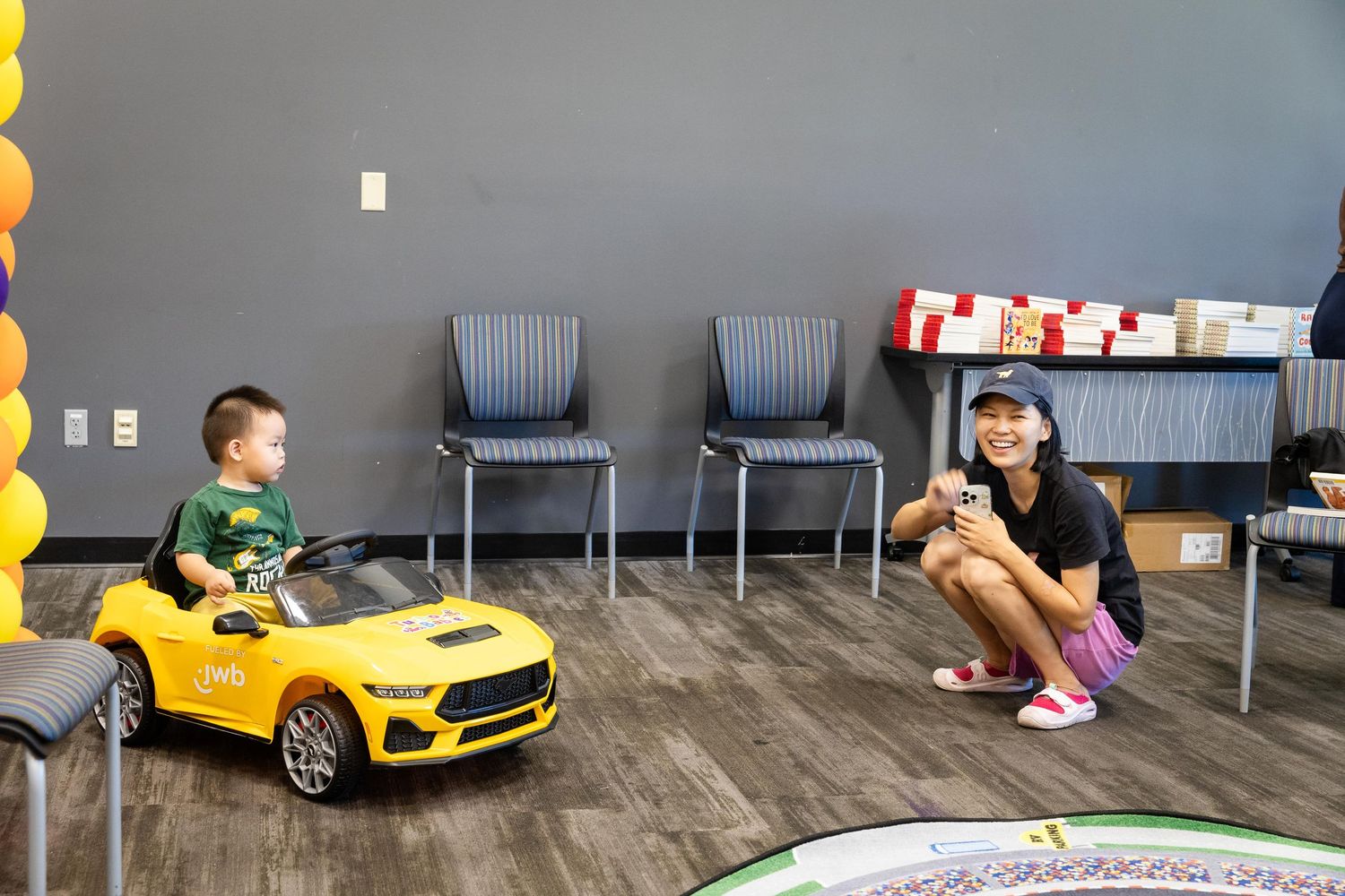 A young toddler rides a yellow toy electric car while a woman crouches nearby smiling in a modern indoor space.