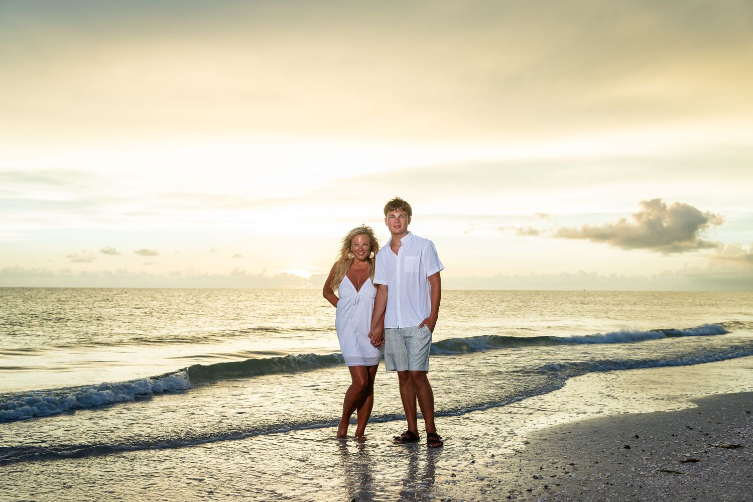 Couple standing together on a sandy beach at sunset, wearing white outfits with ocean waves in the background.