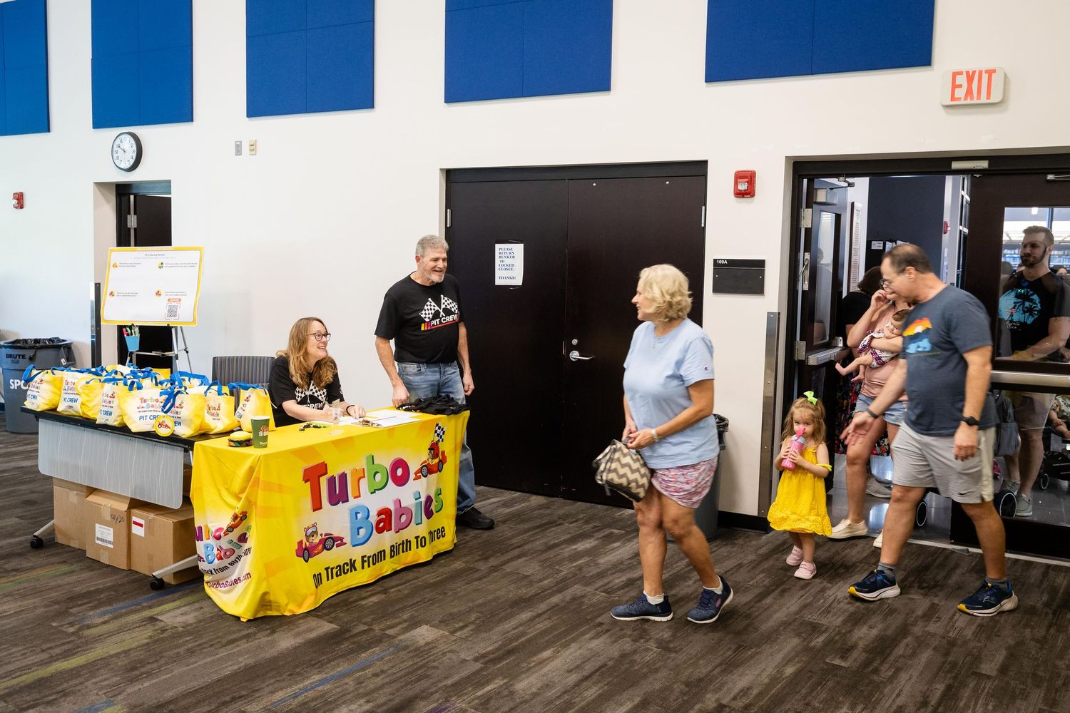 Turbo Babies event booth with staff greeting families in a bright community center near exit doors.