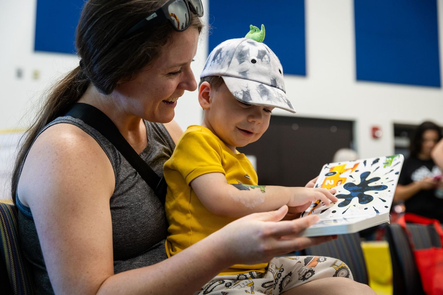 A woman and young boy in a dinosaur cap read a colorful picture book together indoors.
