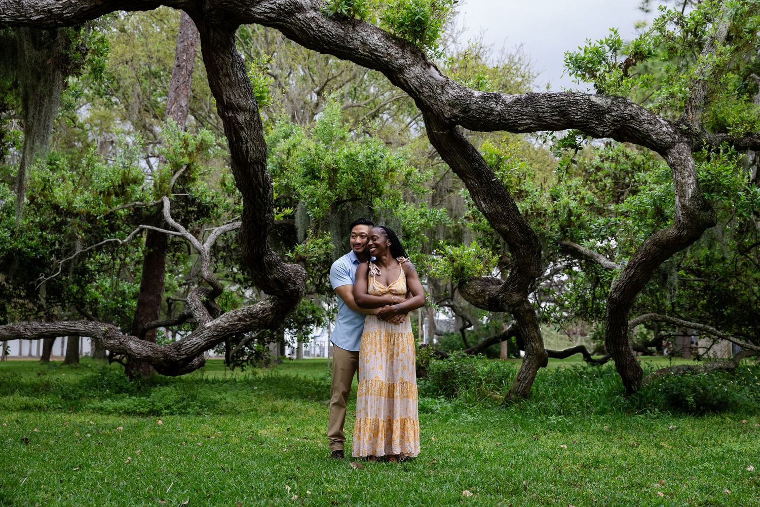 Couple embracing and smiling under sprawling tree branches in lush green park setting.
