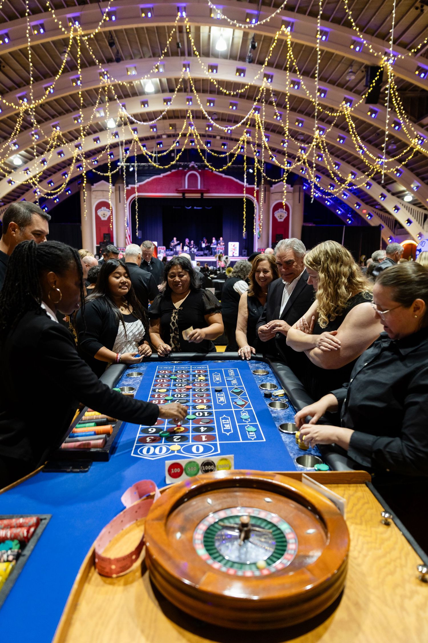 Guests gather around a roulette table at a glamorous casino night event in a decorated ballroom.