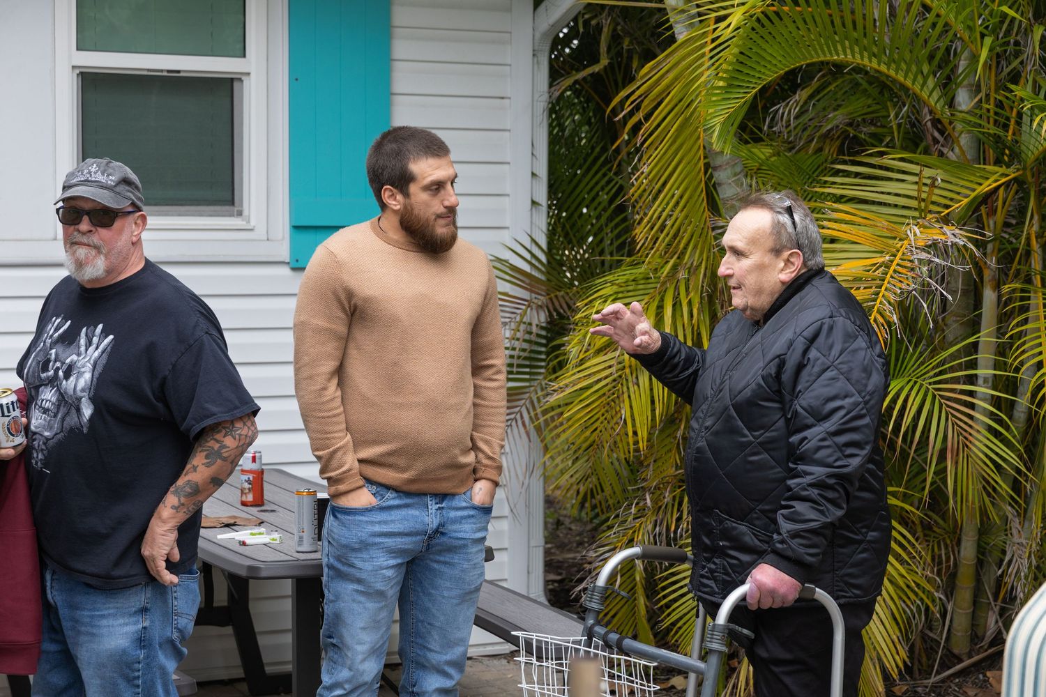 Three men stand outside a house near tropical plants, with an older man using a walker gesturing to two younger men.