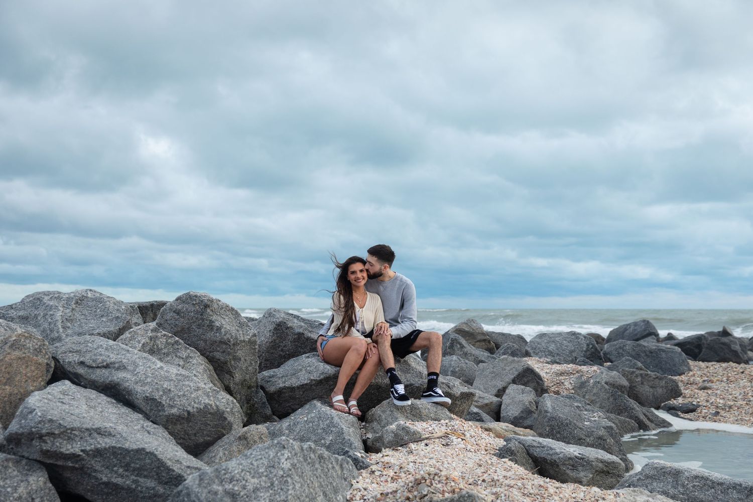 Romantic couple sitting together on rocky beach under cloudy sky by the ocean.