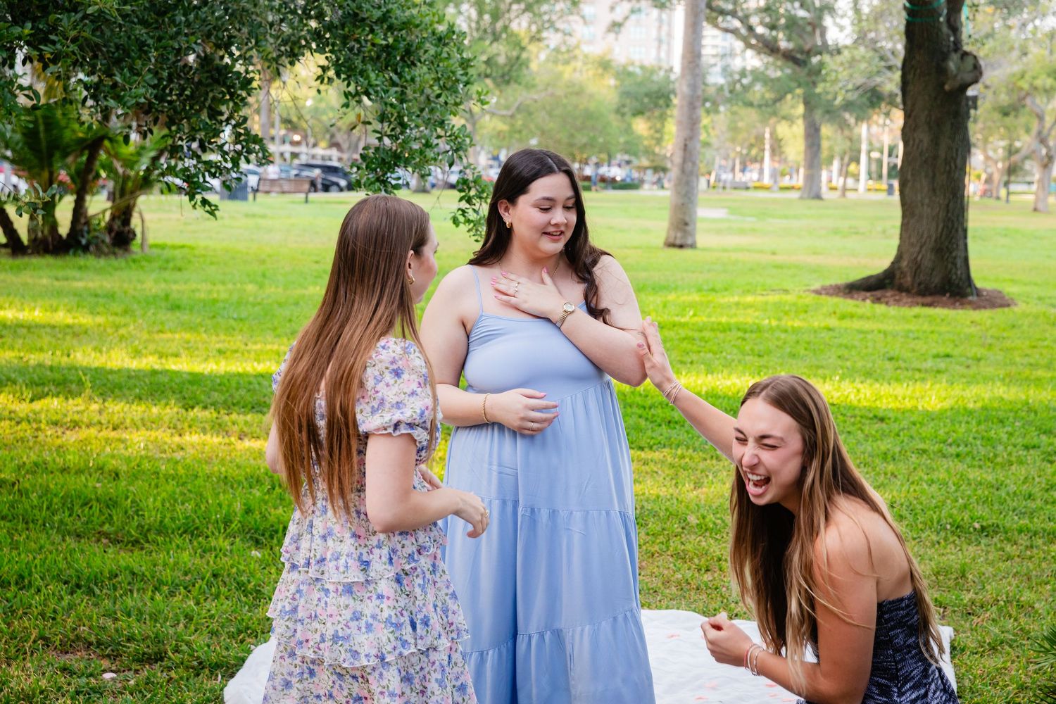 Two friends excitedly react to a pregnant woman in a blue dress at a park maternity photoshoot.