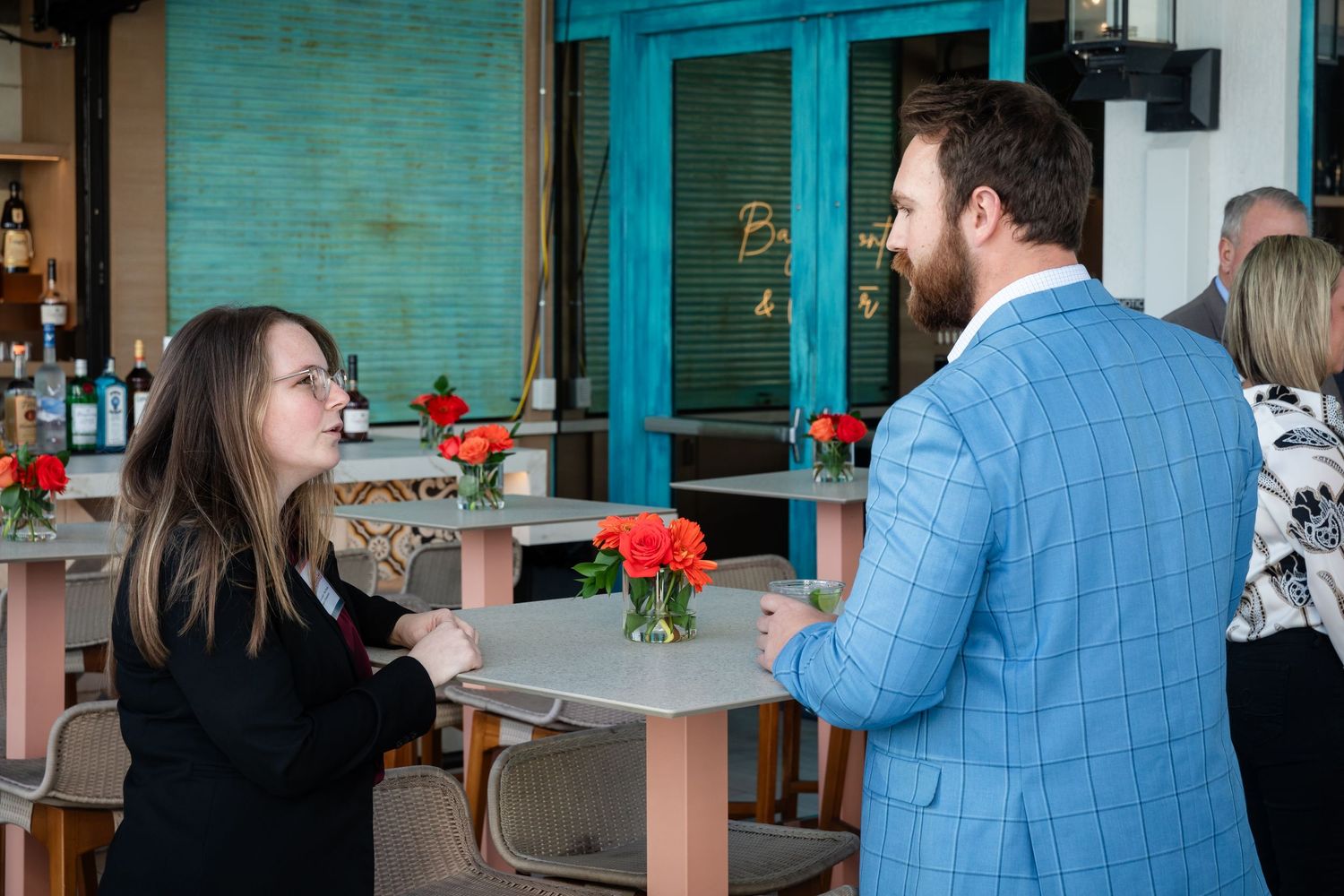 A woman in black and a bearded man in a blue suit converse at an outdoor restaurant with red floral centerpieces.