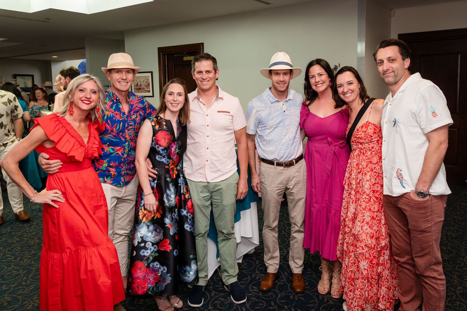 Group of eight smiling adults posing together at a festive indoor gathering, dressed in colorful summer attire.