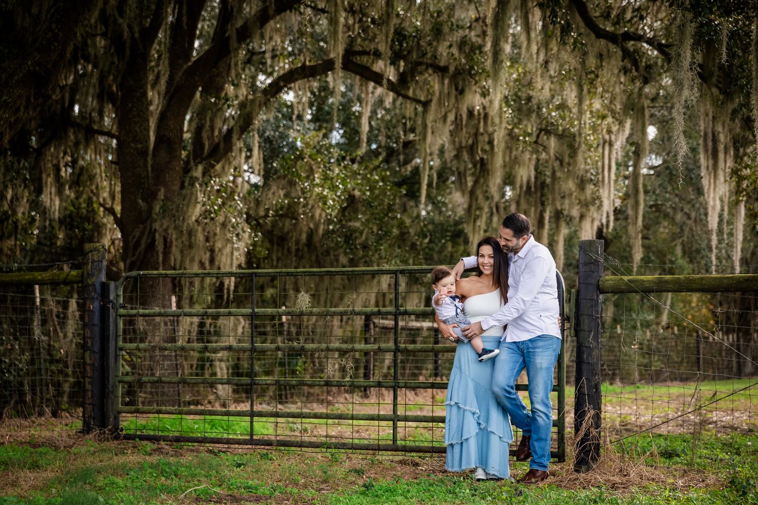 Family portrait with parents holding baby by iron gate under Spanish moss-draped oak trees in lush green outdoor setting.
