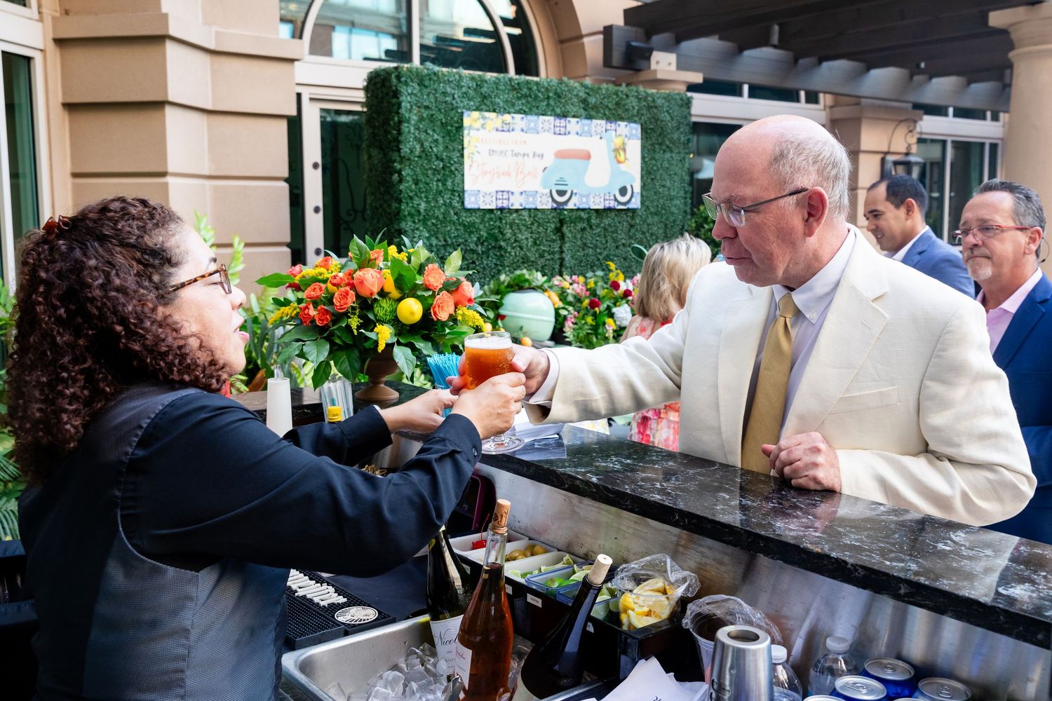 Man in white blazer receiving a cocktail from a bartender at an outdoor bar during a social event.