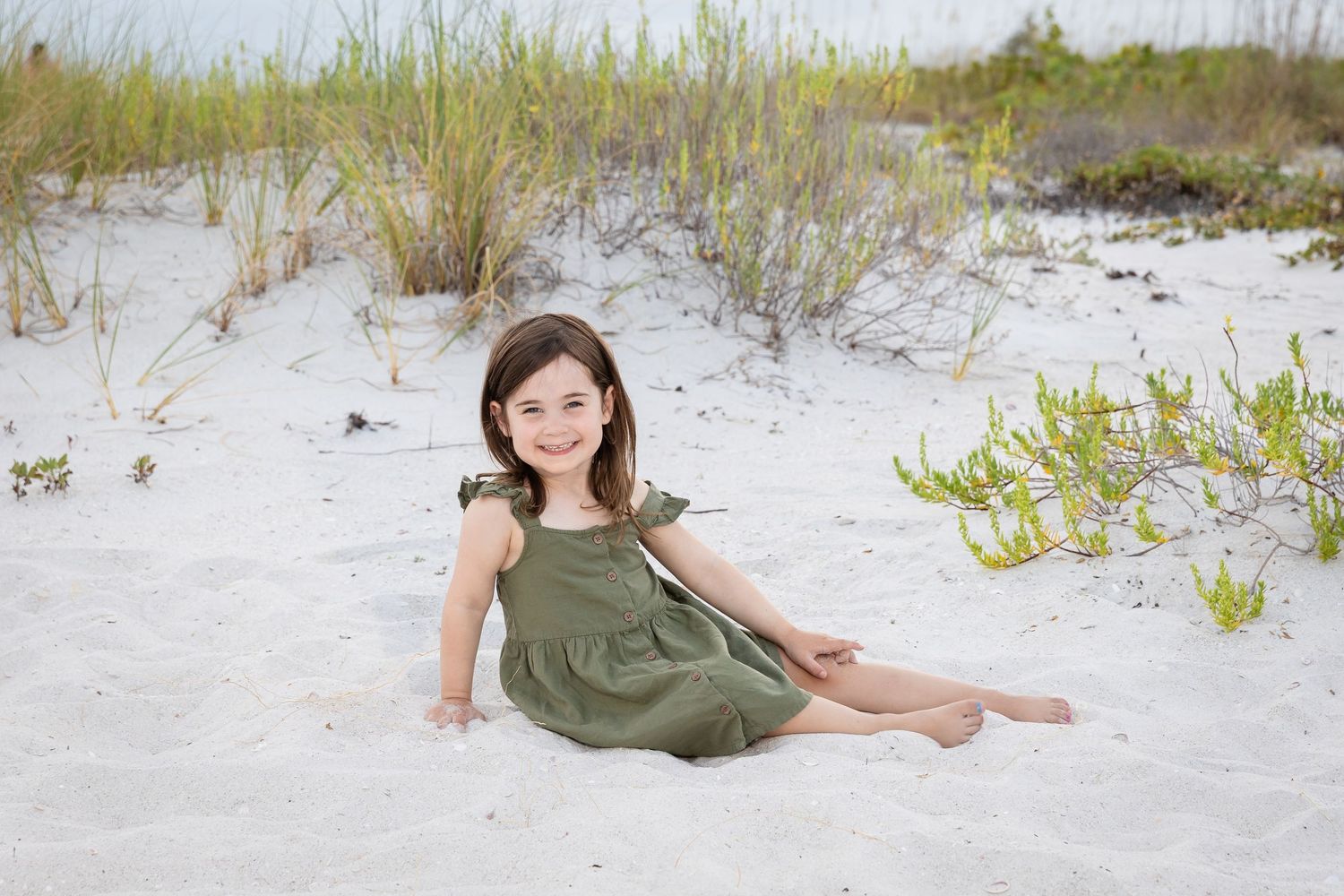 A smiling young girl in a green dress sits on white sand beach dunes with coastal vegetation in the background.