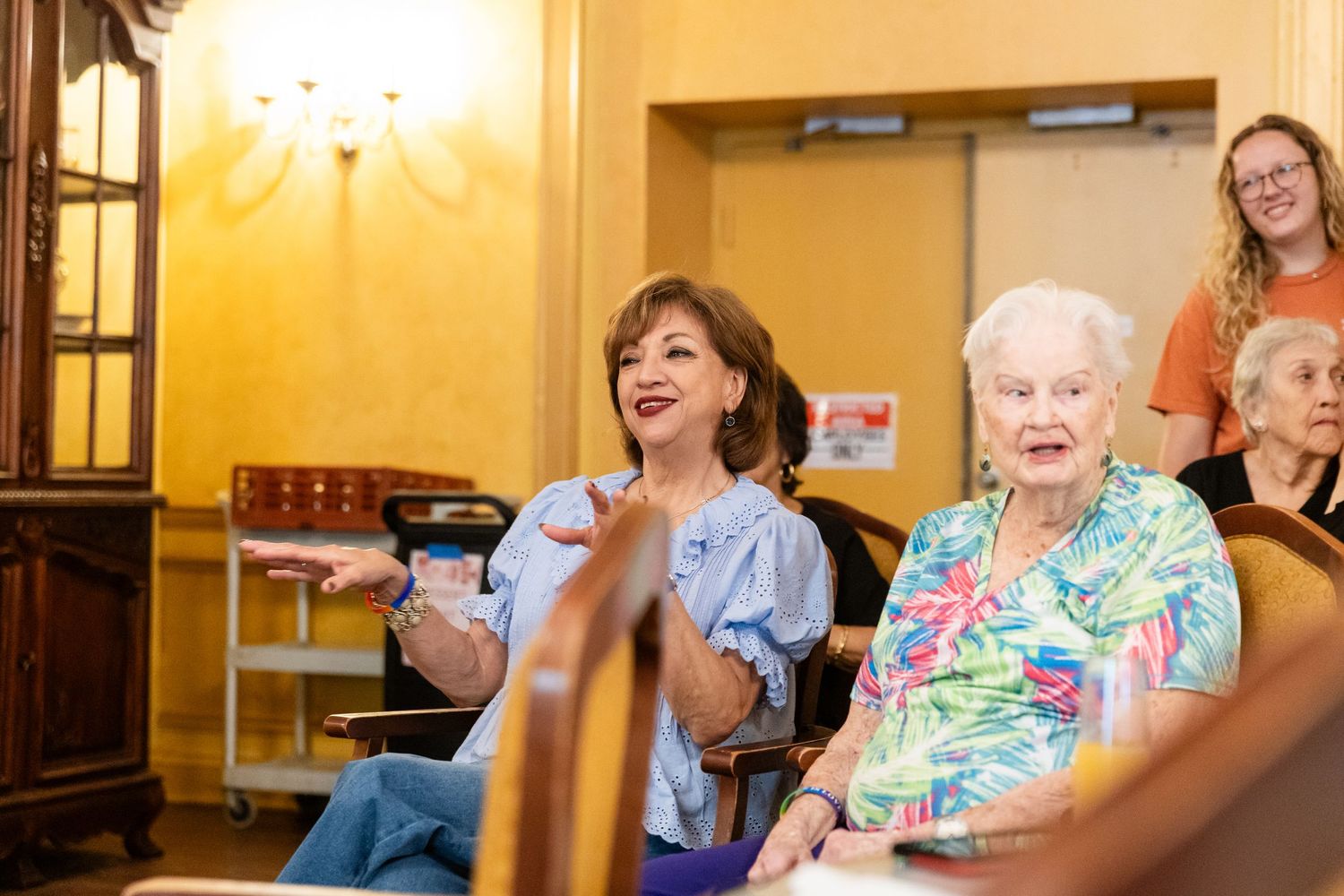Two older women seated at an indoor event, one gesturing expressively while smiling warmly.