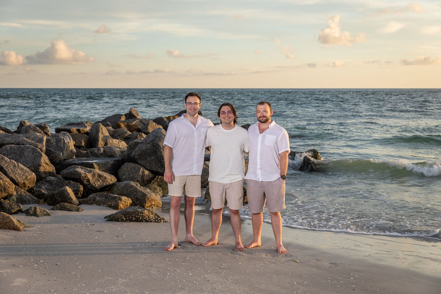 Three men in white shirts and khaki shorts smiling on a beach at sunset near rocky jetty.