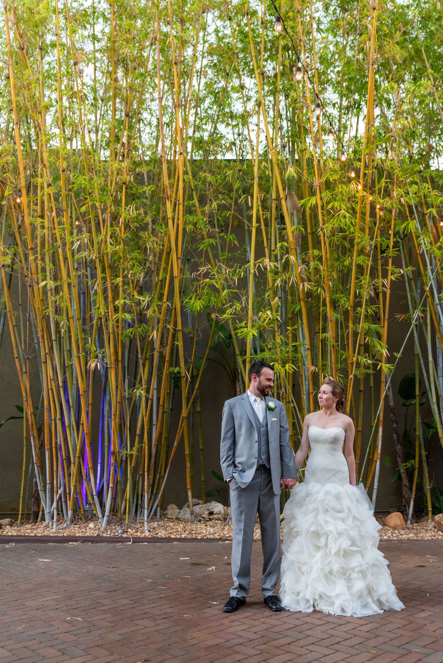 Bride in ruffled gown and groom in gray suit hold hands before tall bamboo backdrop at wedding venue.