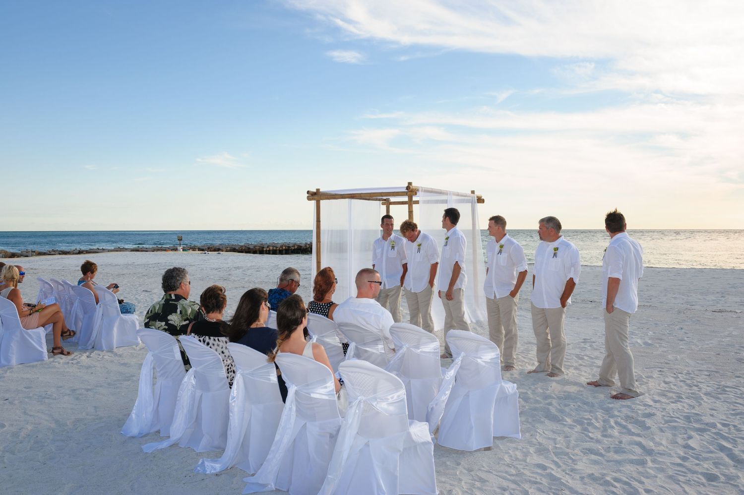 Beach wedding ceremony at sunset with guests seated in white chair covers and groomsmen standing near a bamboo arch.