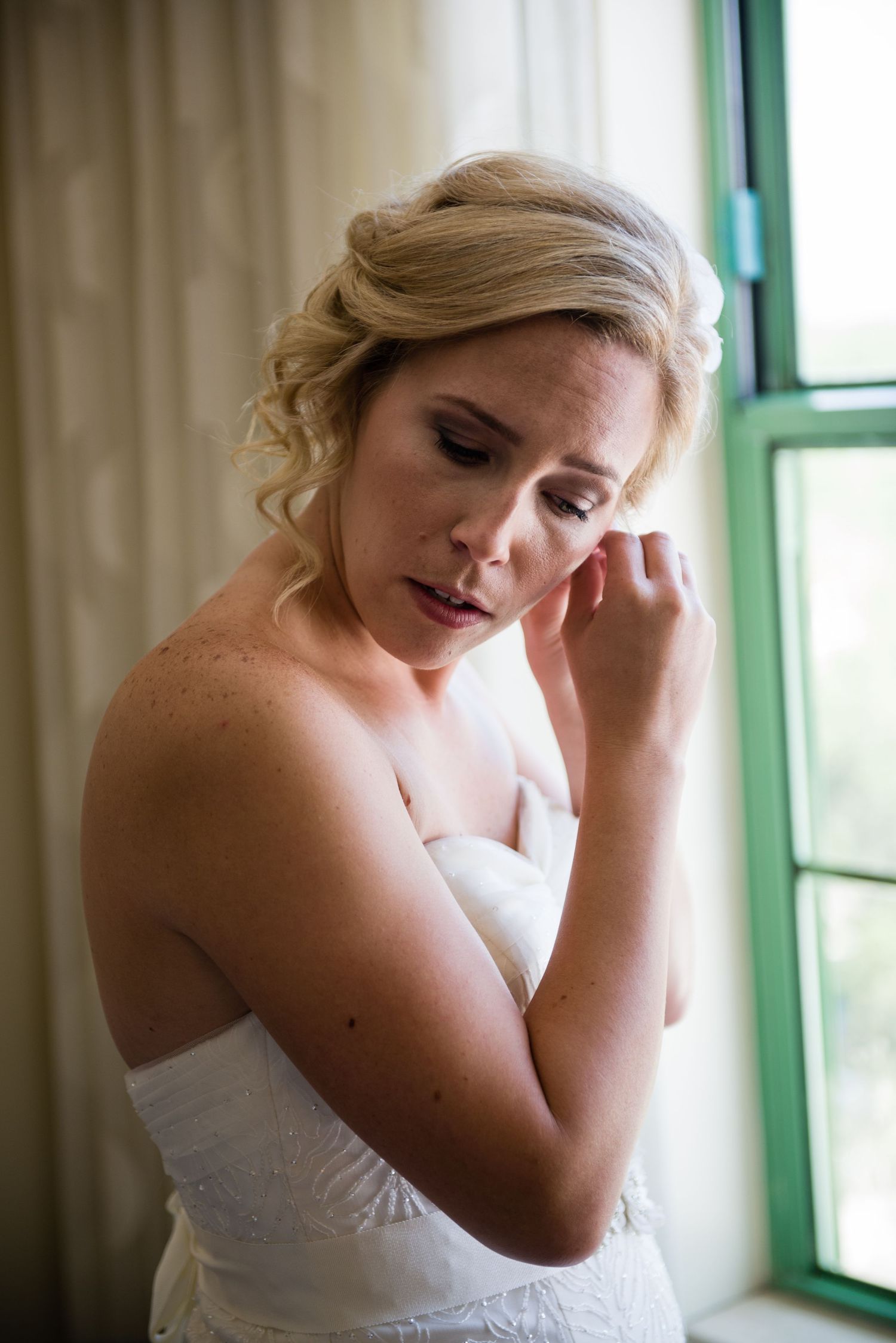 Bride in strapless white wedding dress adjusting earring by window with soft natural light.