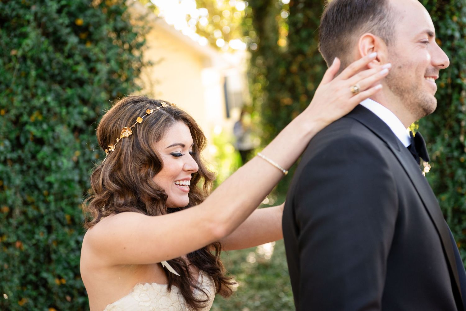 Bride smiling and adjusting groom's collar outdoors at wedding with lush green hedge backdrop.