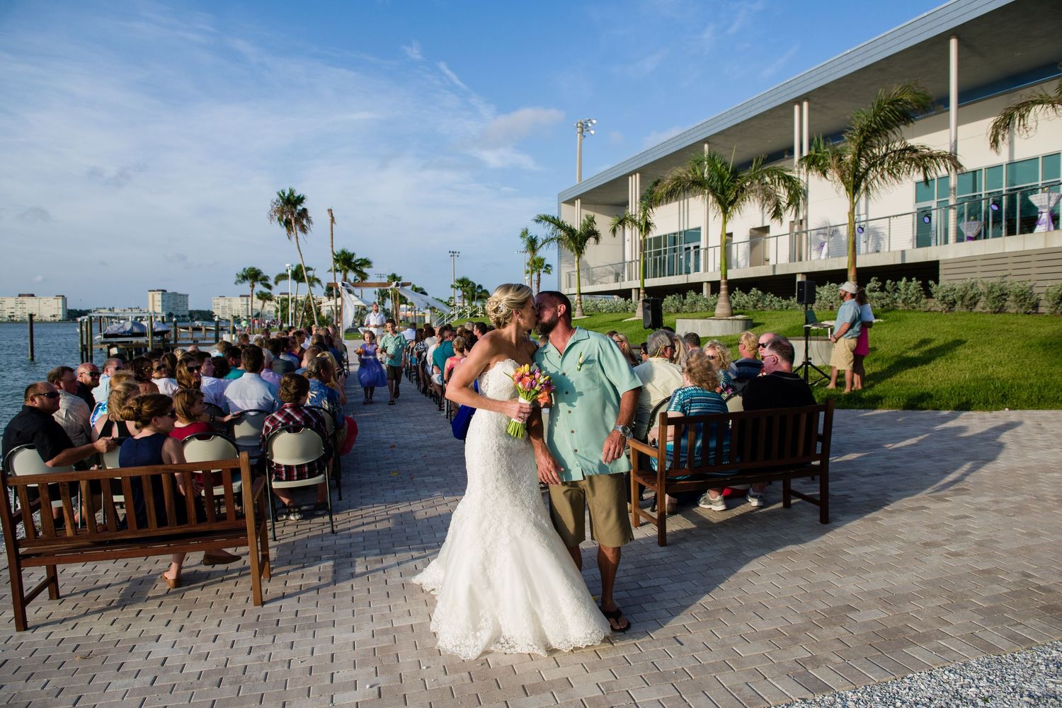 Bride and groom share a kiss during outdoor waterfront wedding ceremony with guests seated on benches near palm trees.