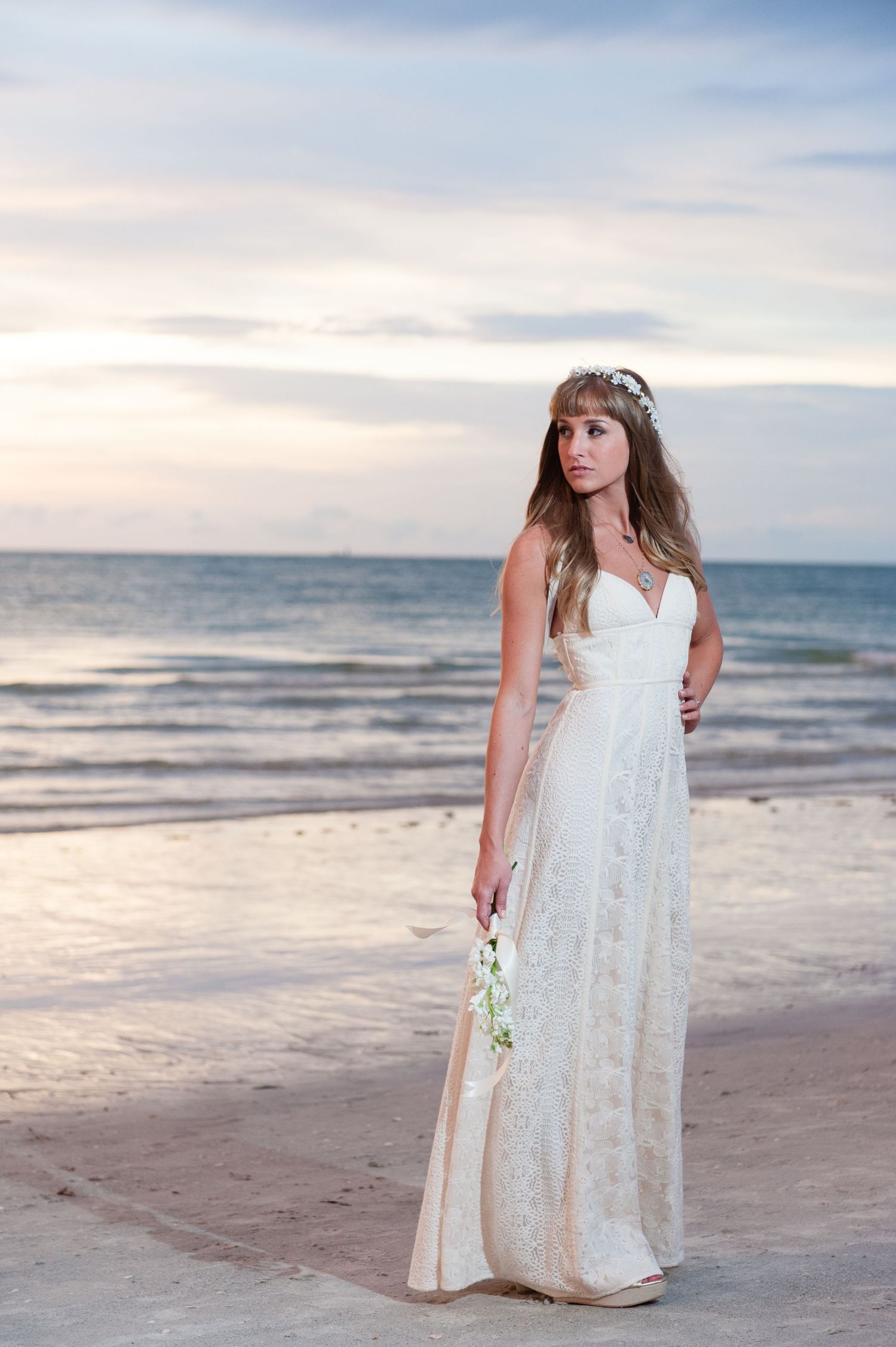 Bride in elegant white lace gown holding bouquet on sandy beach at sunset, wearing delicate floral hair crown.