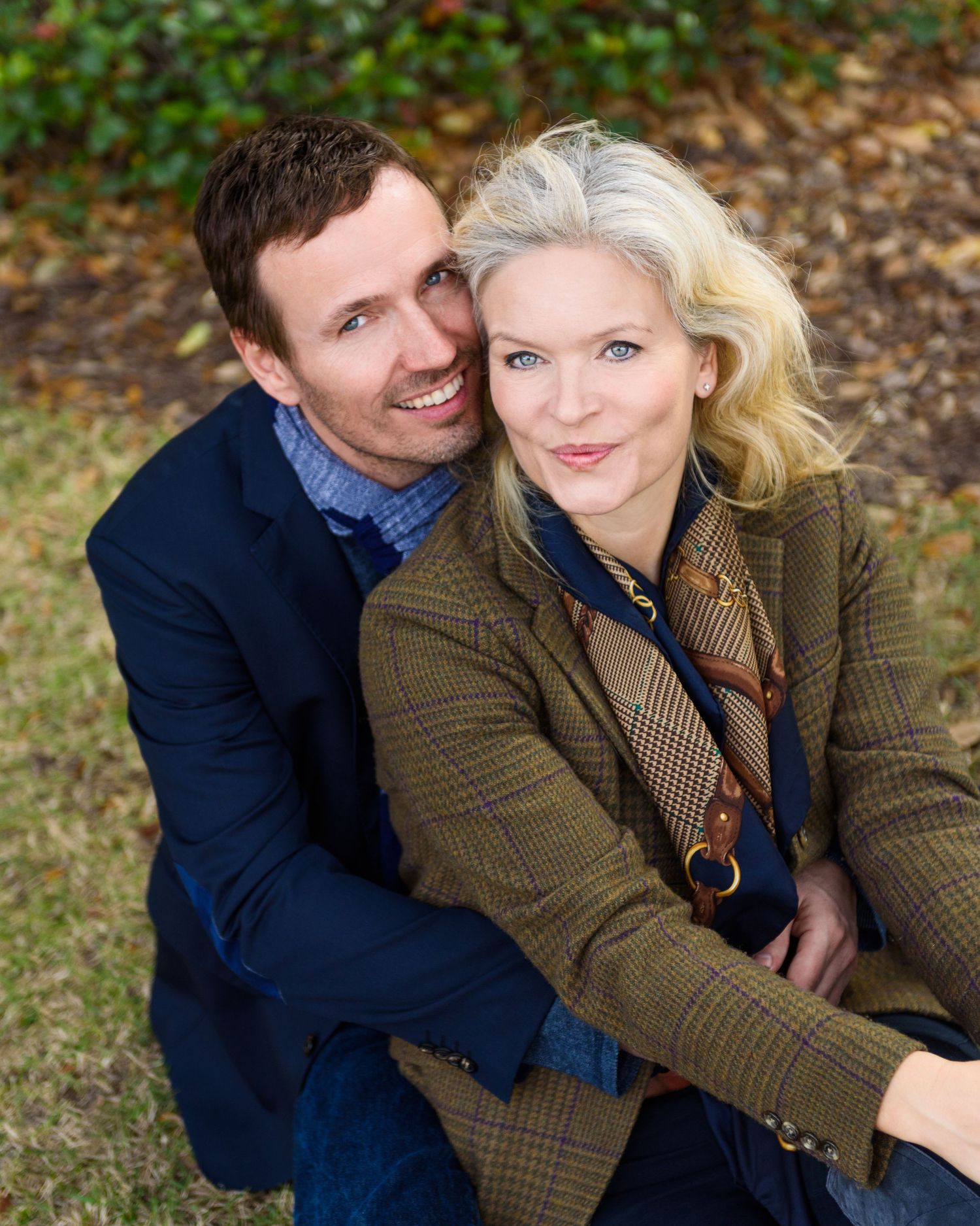 A smiling couple poses outdoors in autumn, wearing smart casual jackets, embracing warmly against a green lawn backdrop.