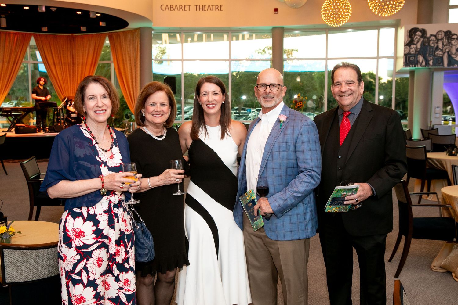 Five elegantly dressed guests smile together at a Cabaret Theatre event, holding drinks and gifts.