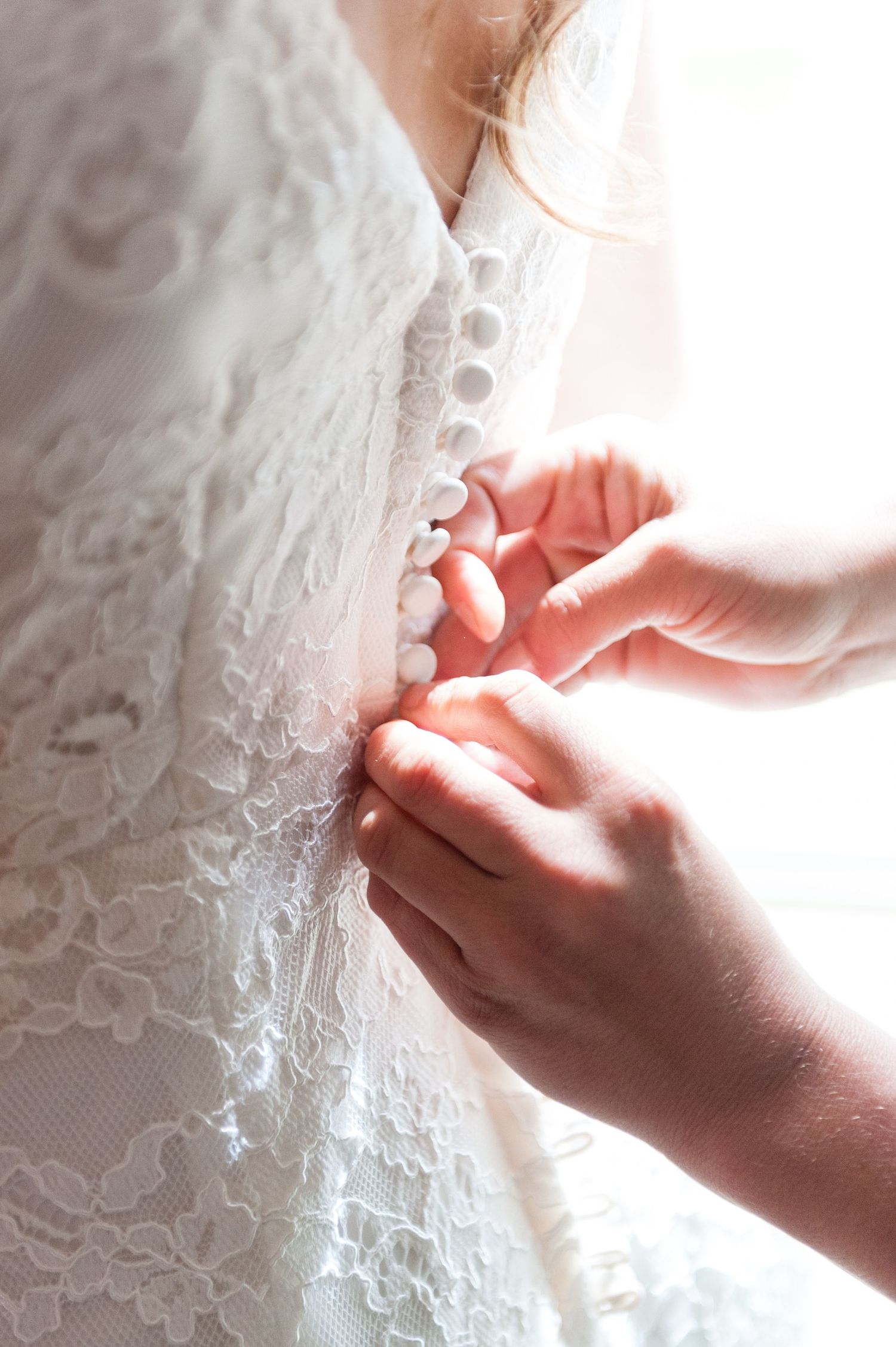 Hands fastening small buttons on the back of a white lace wedding dress.