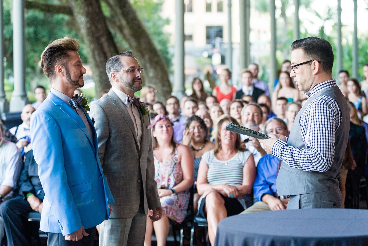 Two grooms exchange vows outdoors at a wedding ceremony with guests seated in the background.