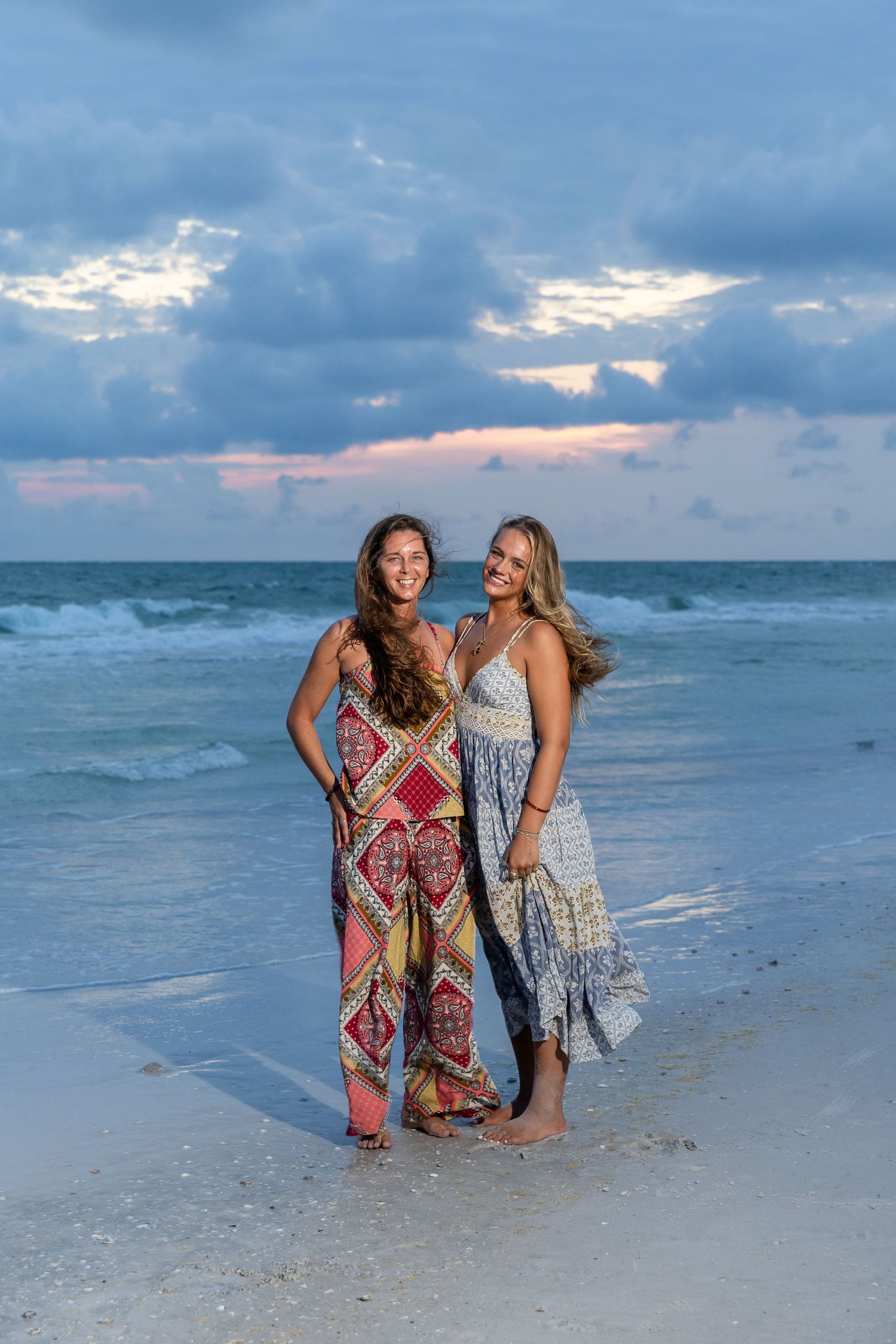 Two women in boho dresses smiling on a sandy beach at sunset with ocean waves and dramatic cloudy sky.