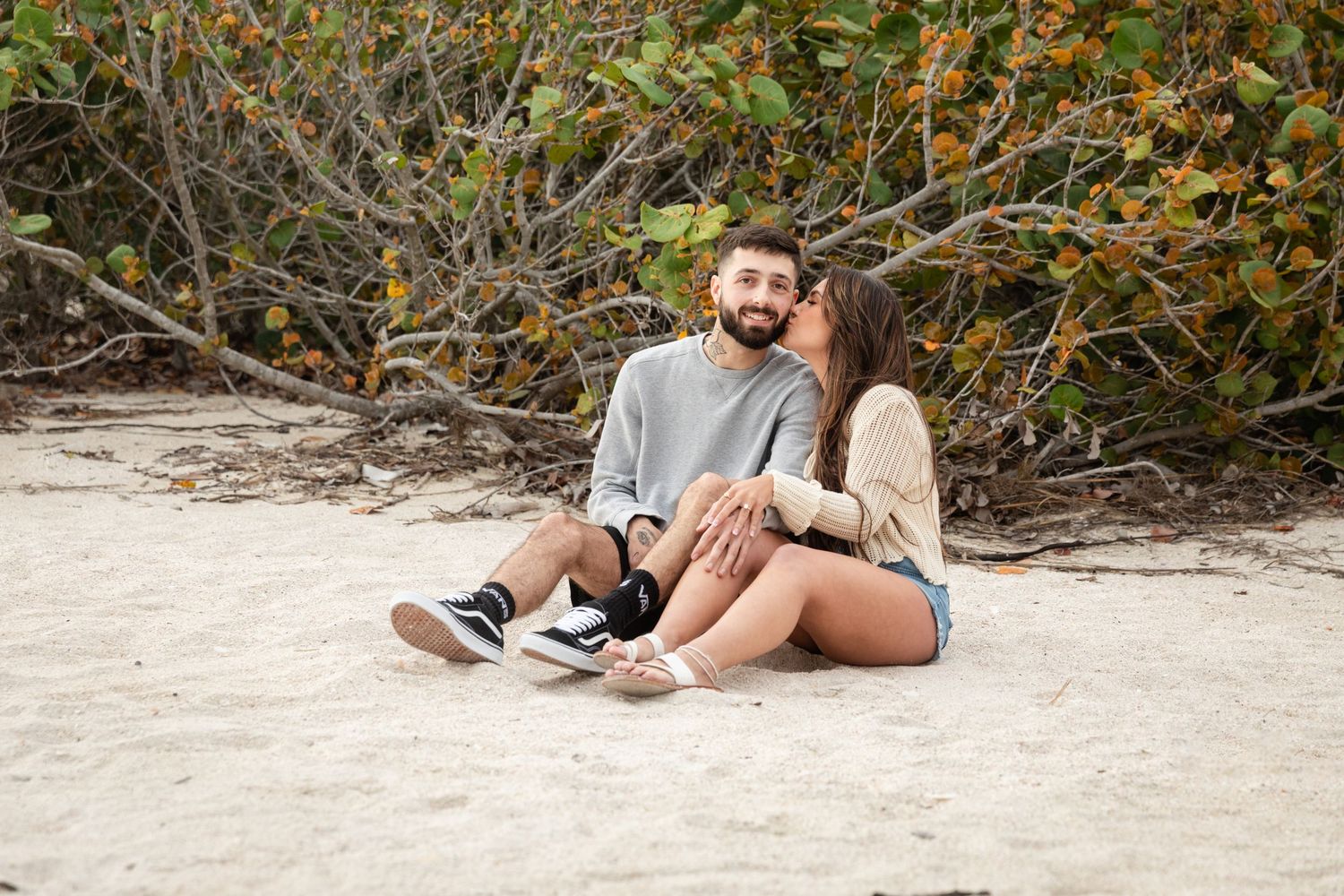 Couple sitting together on white sand beach, leaning against tropical shrubs, smiling and enjoying a romantic outdoor moment.