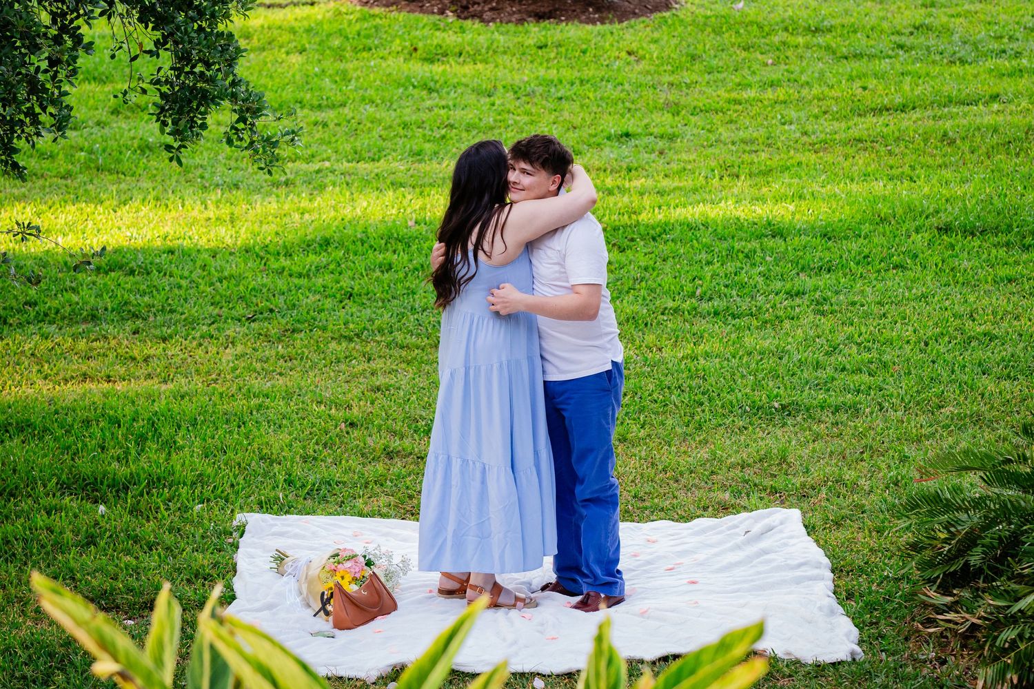 Couple embracing on a white blanket in a lush green park during an outdoor picnic engagement session.