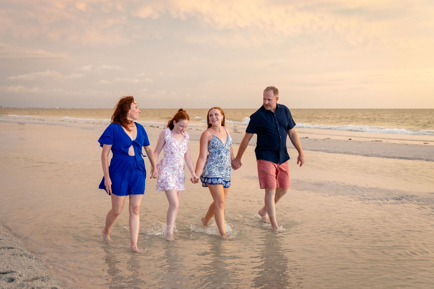Family of four walking hand-in-hand on a beach at sunset, wearing colorful summer outfits.