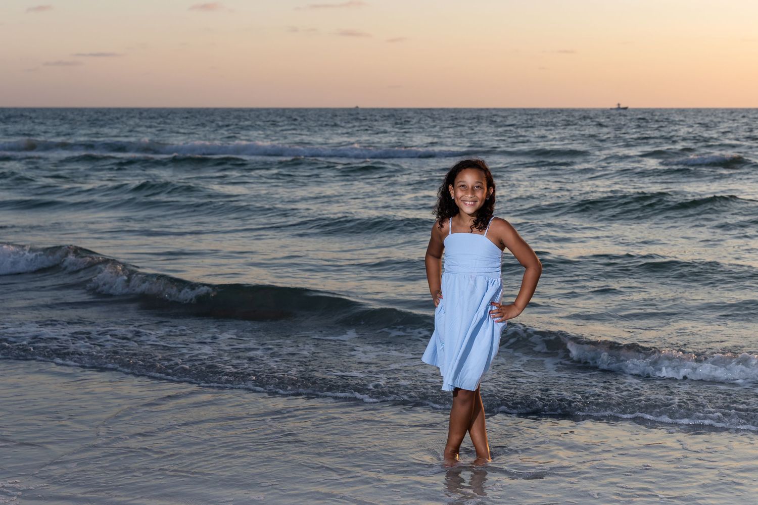 Young girl in blue dress posing on a sandy beach at sunset with ocean waves in the background.