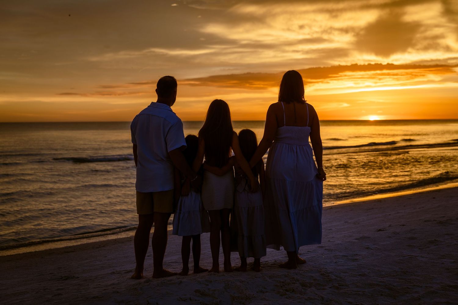 Family of four silhouetted against a stunning golden sunset on a peaceful beach shoreline.
