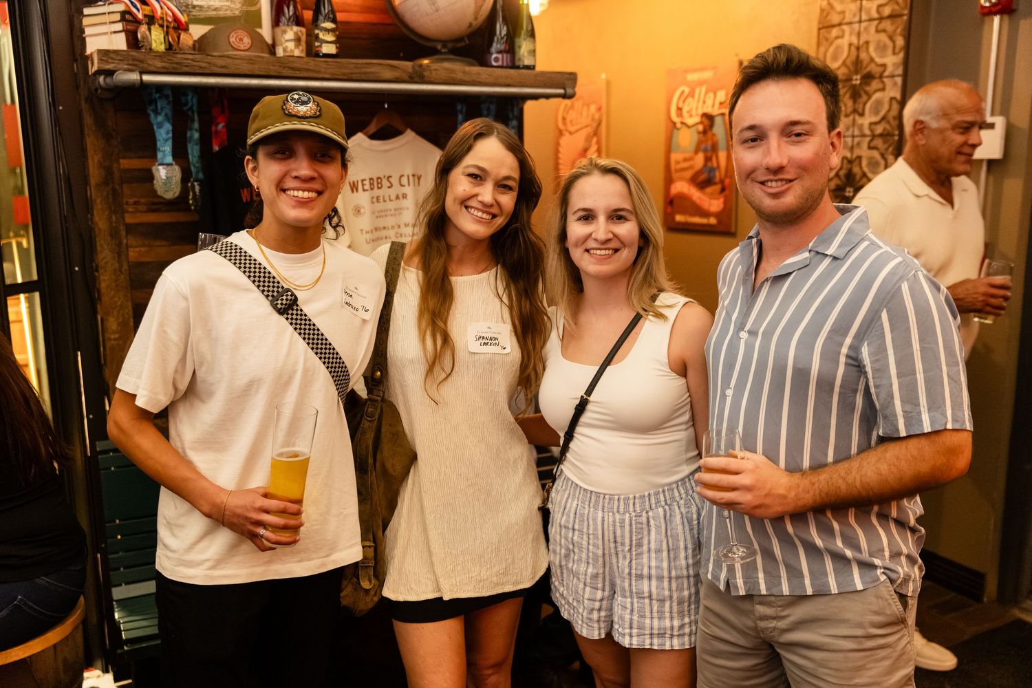 Four young adults smiling together at a lively indoor brewery social event.