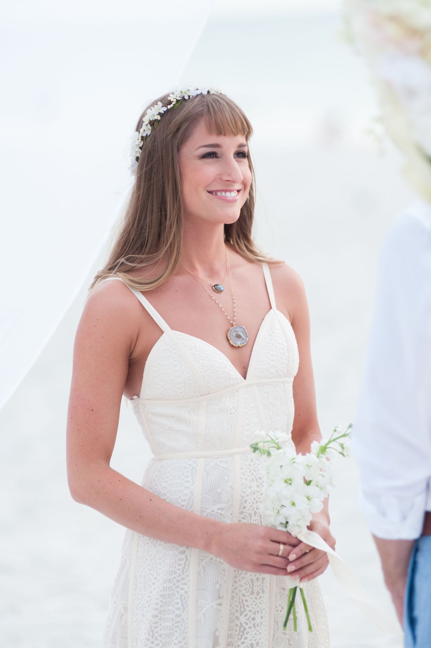 Beautiful bride in lace gown wearing floral crown holds white bouquet while smiling at beach wedding on Treasure Island, Florida.