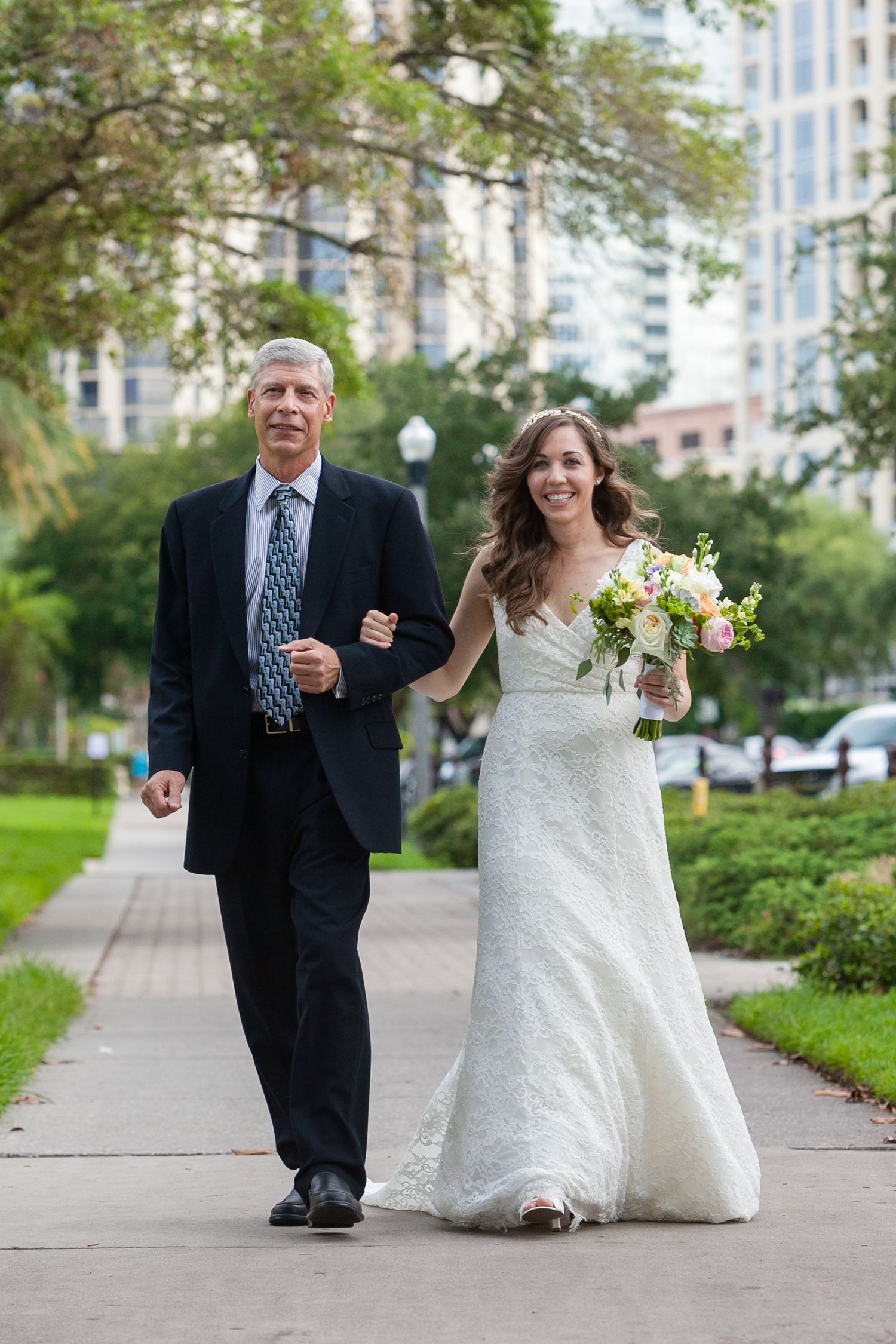 Bride in lace gown walks arm-in-arm with her father down a tree-lined urban sidewalk holding a pastel bouquet Museum of Fine Arts, St. Pete.