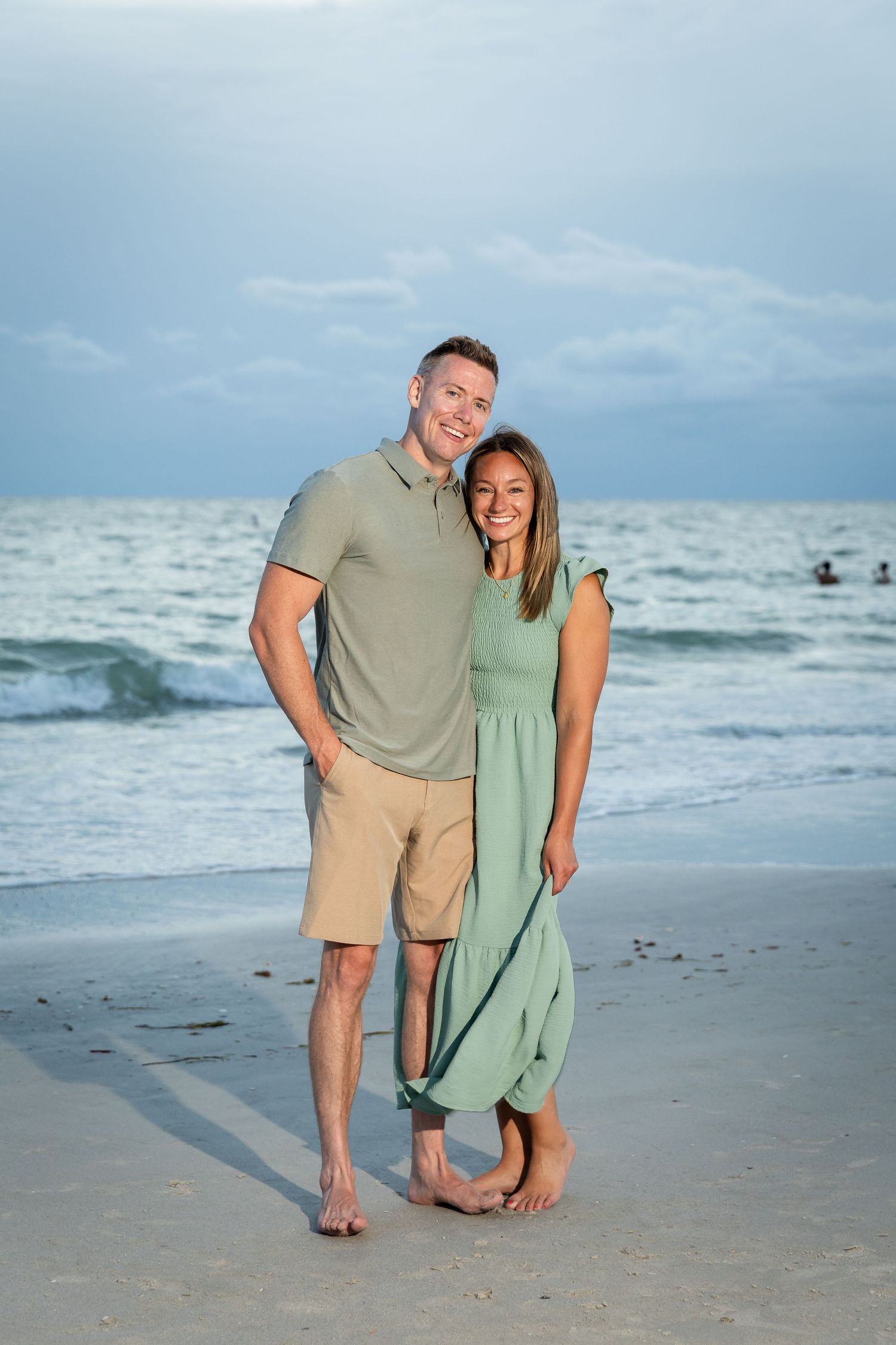 Smiling couple in coordinating green outfits standing barefoot together on sandy beach at dusk.