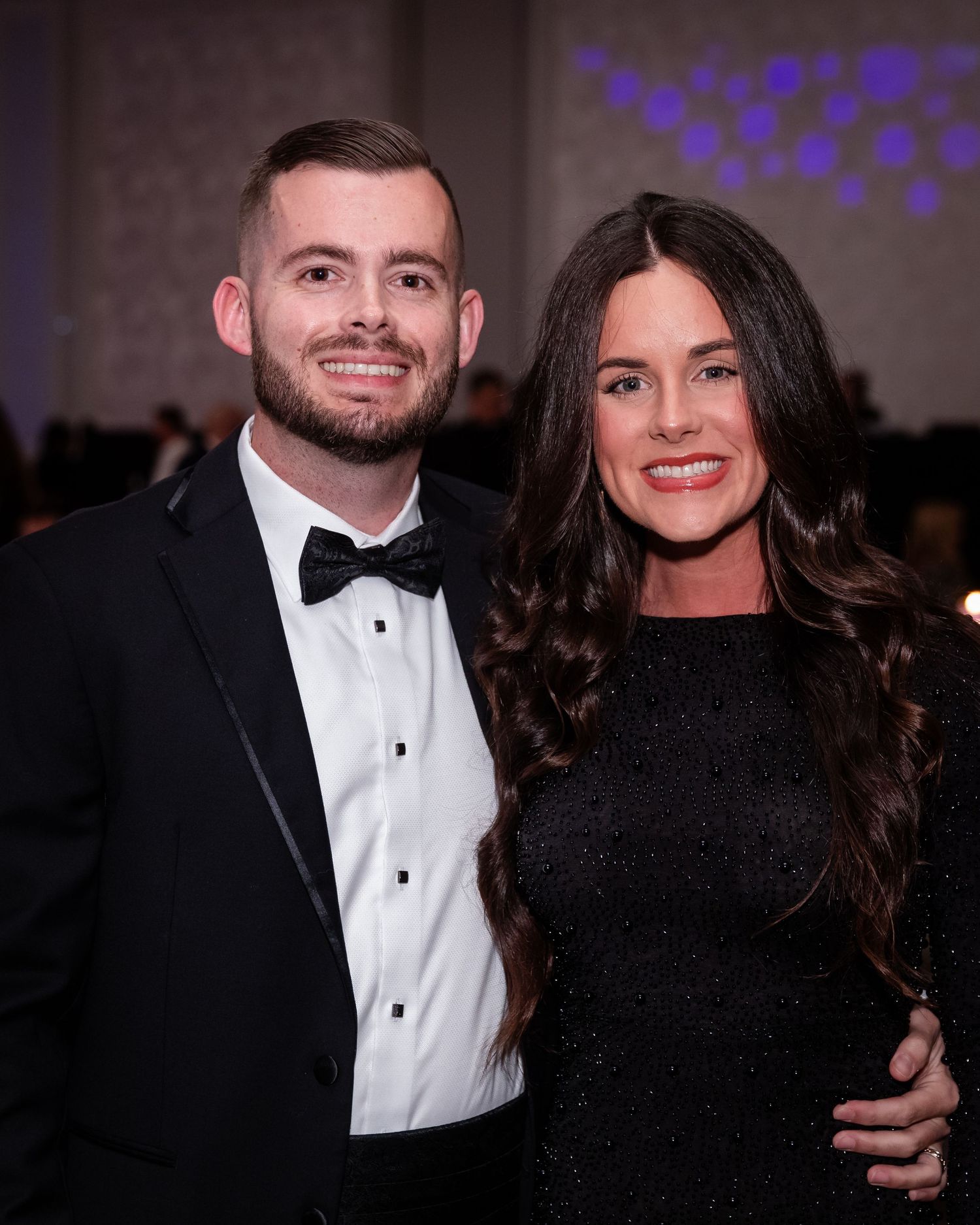 Smiling couple posing at formal event, man in tuxedo with bow tie and woman in black dress.