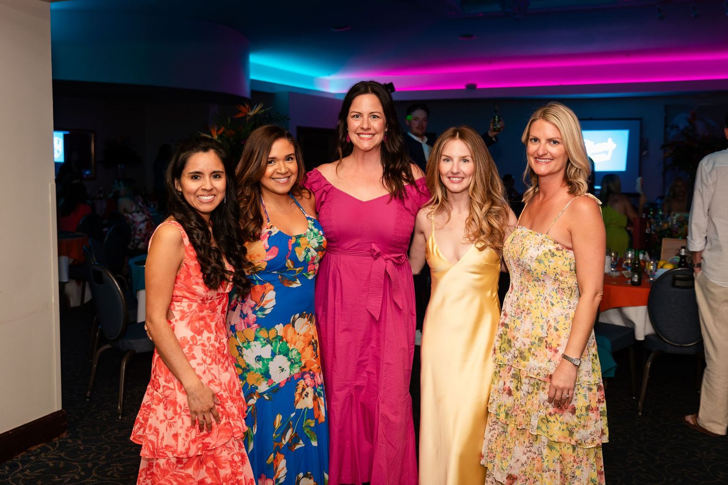 Five women in colorful formal dresses smile together at an elegant indoor event with purple lighting.