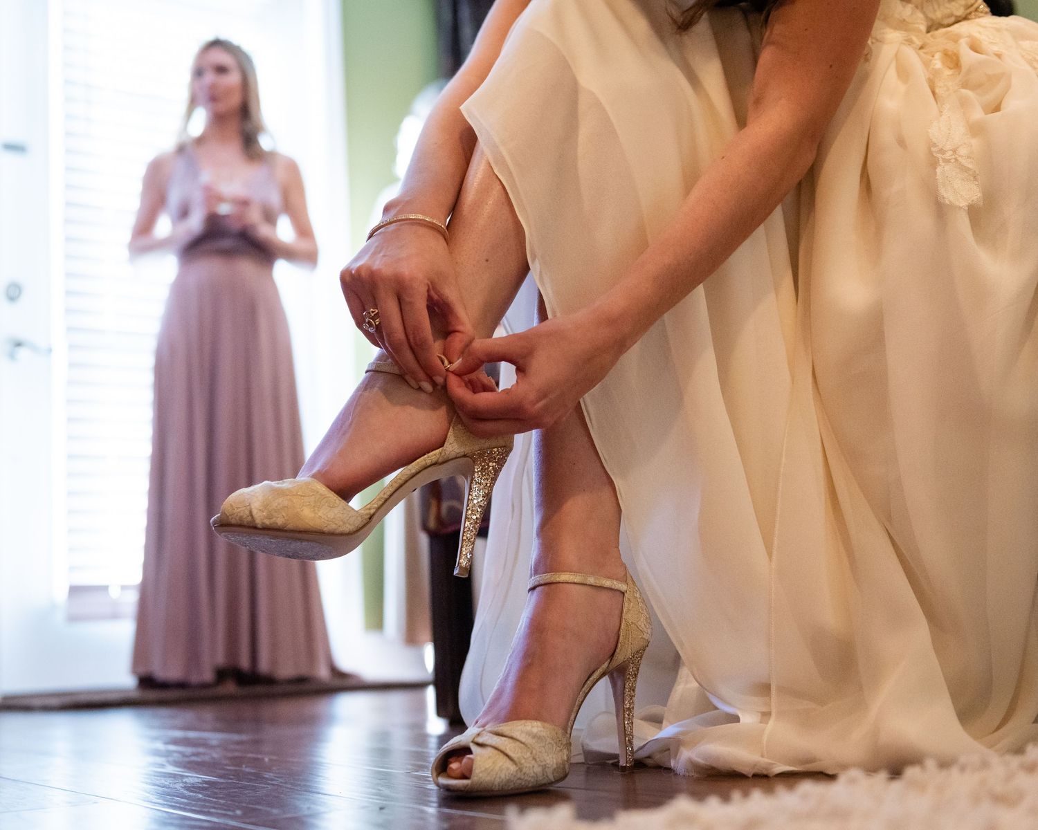 Bride in white gown fastening strappy gold heels while bridesmaid in mauve dress stands in background.