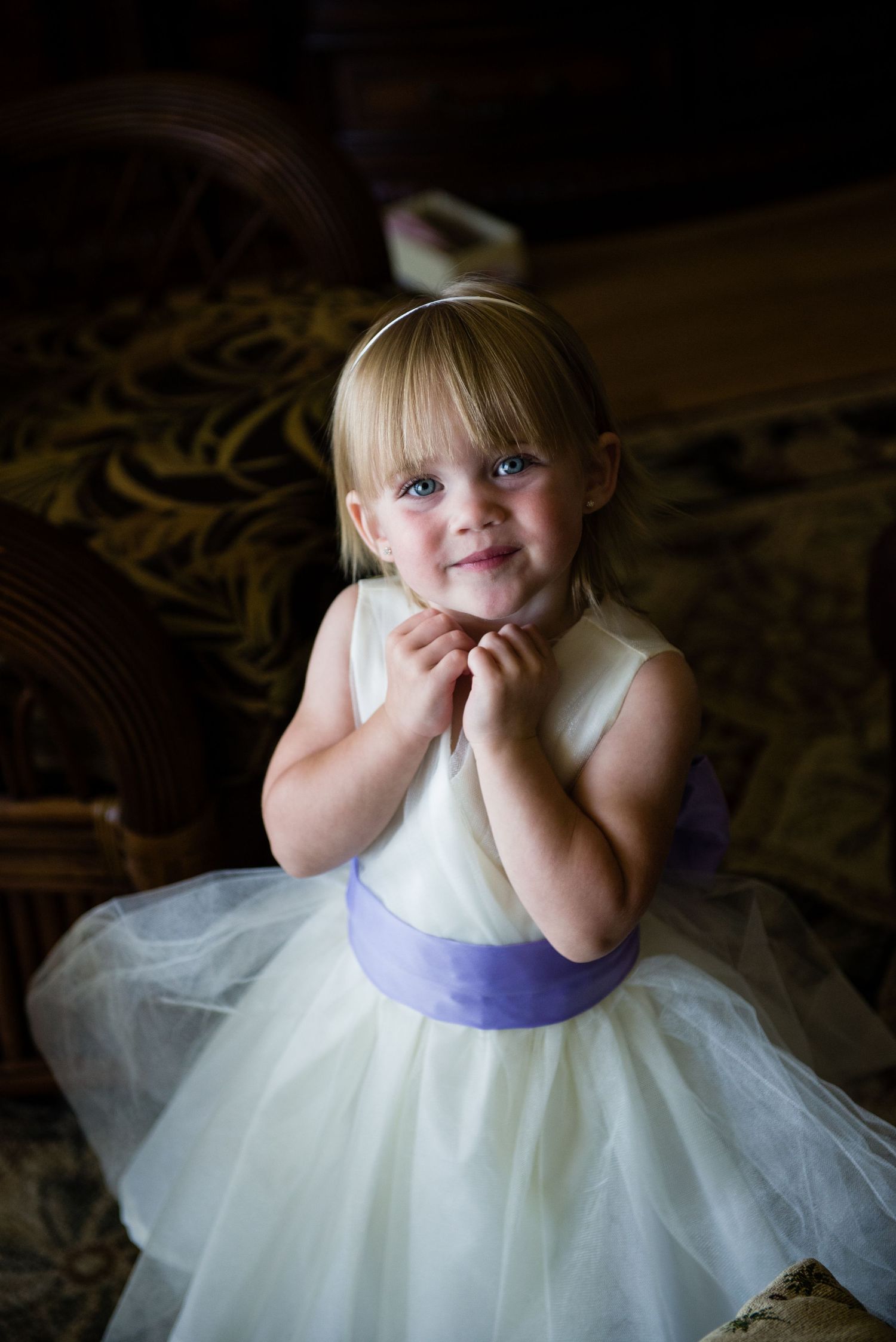 Adorable flower girl in white and lavender tulle dress with headband smiling sweetly at a wedding.