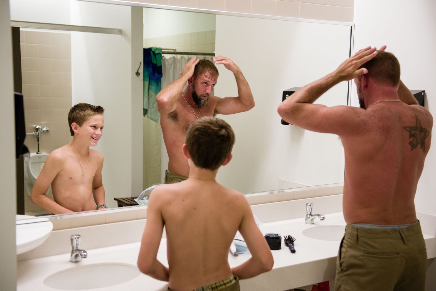 Two shirtless males and a boy fixing their hair in a bright bathroom mirror.