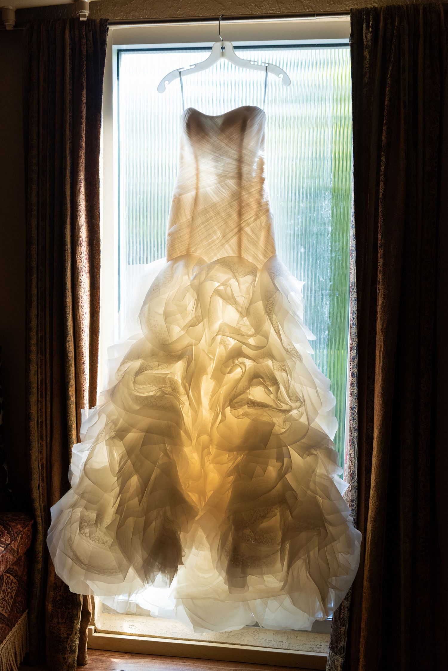 A stunning strapless wedding gown with ruffled skirt hangs backlit in a window, glowing in warm golden light.
