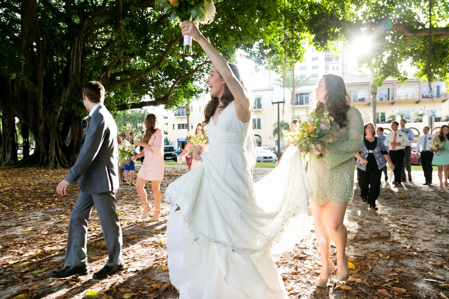 Joyful bride in white gown tosses bouquet outdoors under large tree with wedding guests nearby.
