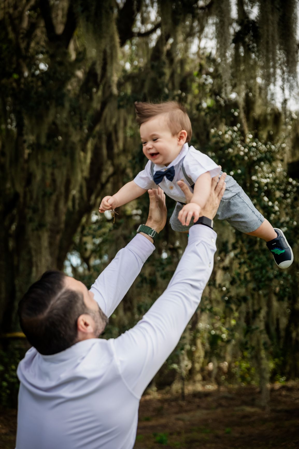Joyful father lifting laughing baby boy high in air under Spanish moss trees during outdoor family photo session.