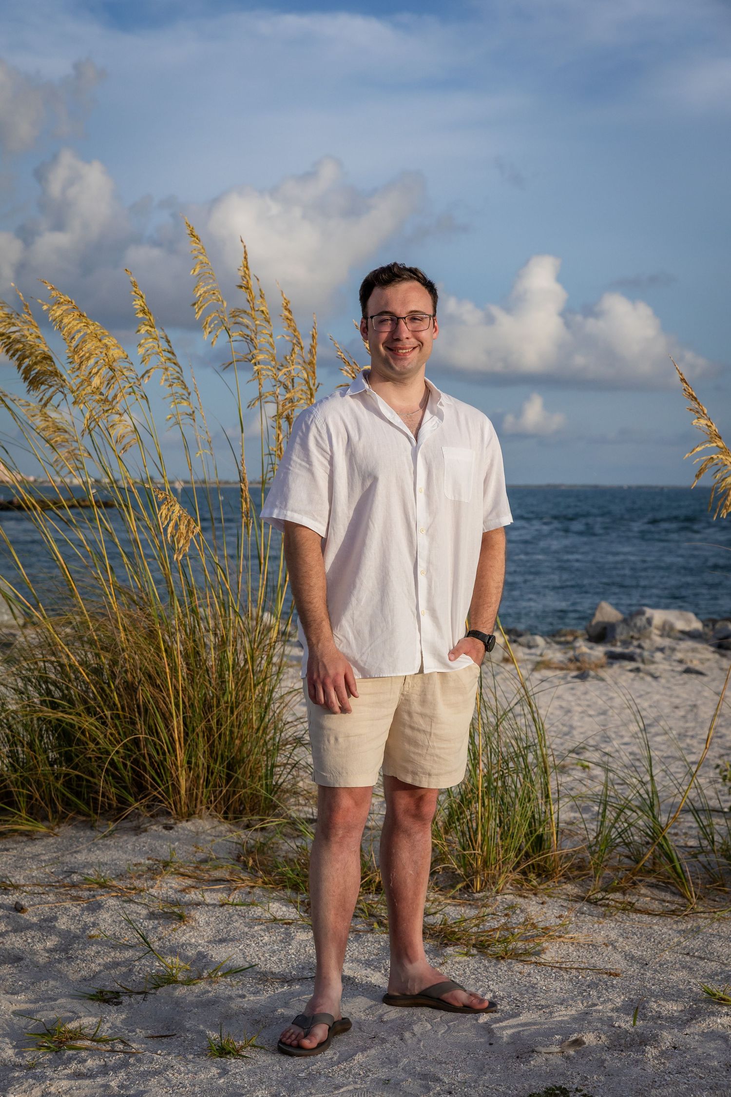 A smiling man in white shirt and khaki shorts stands barefoot on a sandy beach with sea oats and ocean backdrop.