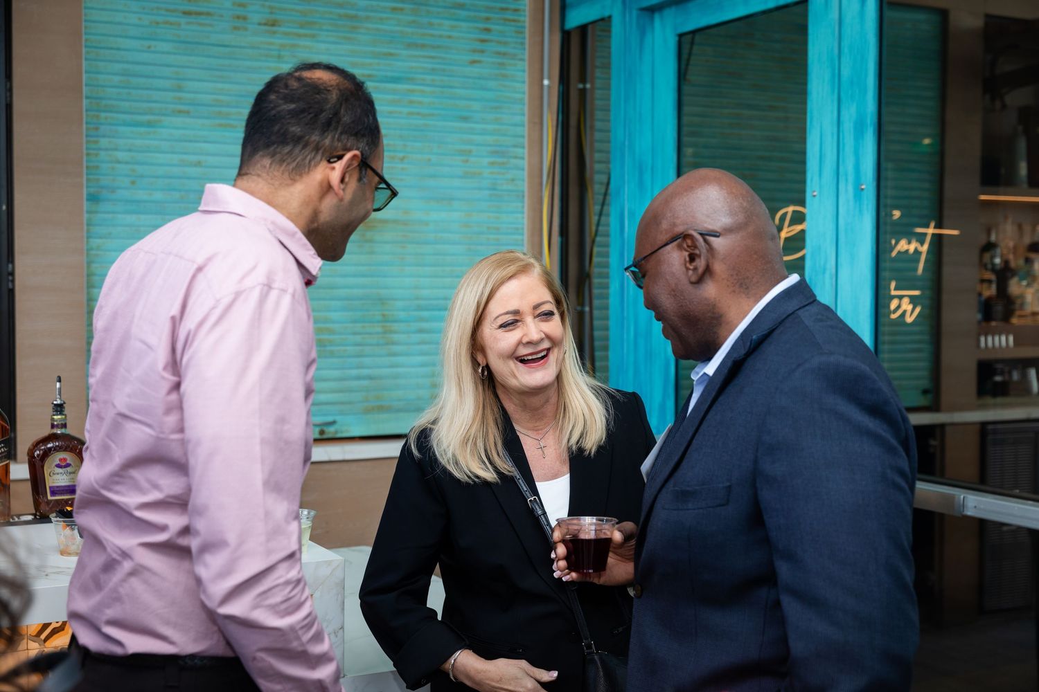 Three business professionals laugh and socialize at an outdoor networking event near a teal-accented venue wall.