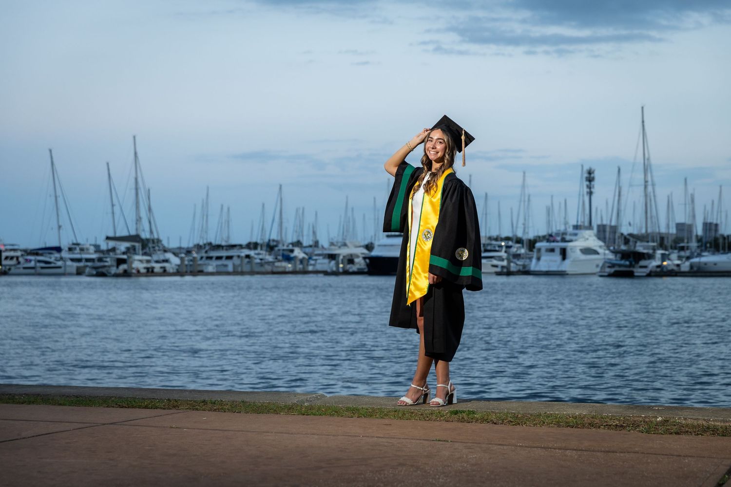 Graduate in black cap and gown with yellow stole poses at marina waterfront during twilight ceremony photos.