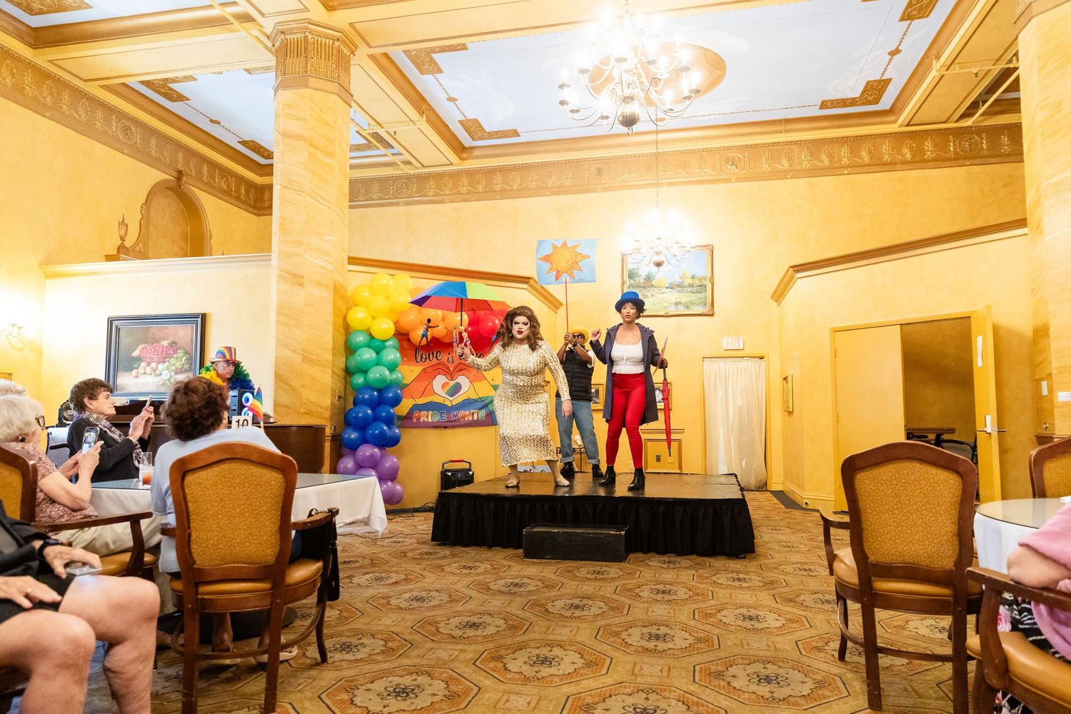 Performers on stage at a colorful Pride event in an elegant ballroom with rainbow decorations and seated audience.